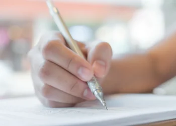Mão segurando caneta prateada escreve em papel com texto impresso em fundo desfocado, simbolizando as inscrições para o concurso Câmara de Vila Boa.