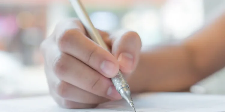 Mão segurando caneta prateada escreve em papel com texto impresso em fundo desfocado, simbolizando as inscrições para o concurso Câmara de Vila Boa.
