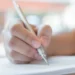 Mão segurando caneta prateada escreve em papel com texto impresso em fundo desfocado, simbolizando as inscrições para o concurso Câmara de Vila Boa.