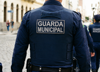 Guarda Municipal de costas com uniforme azul e patch, patrulhando rua de paralelepípedos em grupo, simbolizando o concurso em Caldas Novas/GO.