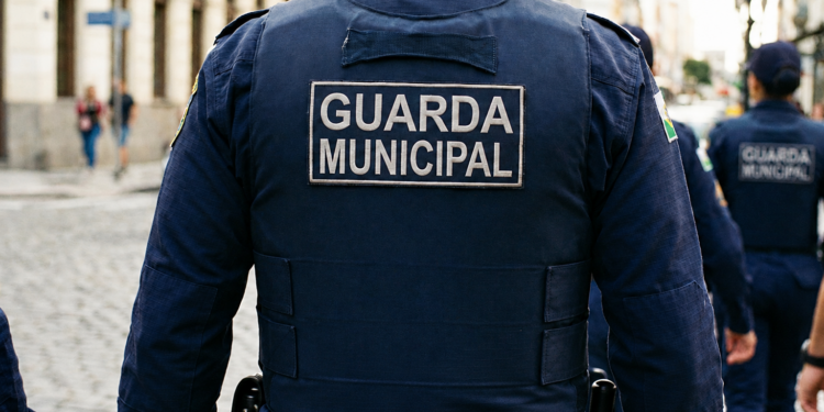 Guarda Municipal de costas com uniforme azul e patch, patrulhando rua de paralelepípedos em grupo, simbolizando o concurso em Caldas Novas/GO.