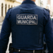 Guarda Municipal de costas com uniforme azul e patch, patrulhando rua de paralelepípedos em grupo, simbolizando o concurso em Caldas Novas/GO.