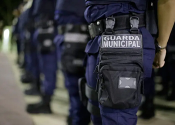 Agentes da Guarda Municipal perfilados de costas, com uniforme azul-escuro e o distintivo da corporação visível no equipamento de cintura.