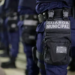 Agentes da Guarda Municipal perfilados de costas, com uniforme azul-escuro e o distintivo da corporação visível no equipamento de cintura.