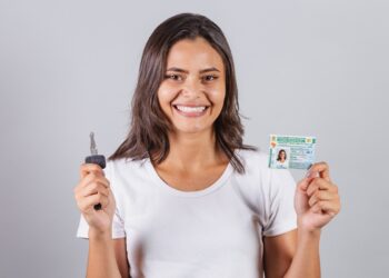 Mulher sorridente mostra CNH brasileira e chave de carro sobre fundo cinza claro, simbolizando os concursos que possuem exigência de CNH.