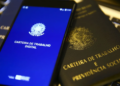 Celular com a tela inicial do aplicativo Carteira de Trabalho Digital apoiado sobre exemplares impressos da Carteira de Trabalho e Previdência Social, simbolizando a consulta do PIS/Pasep.