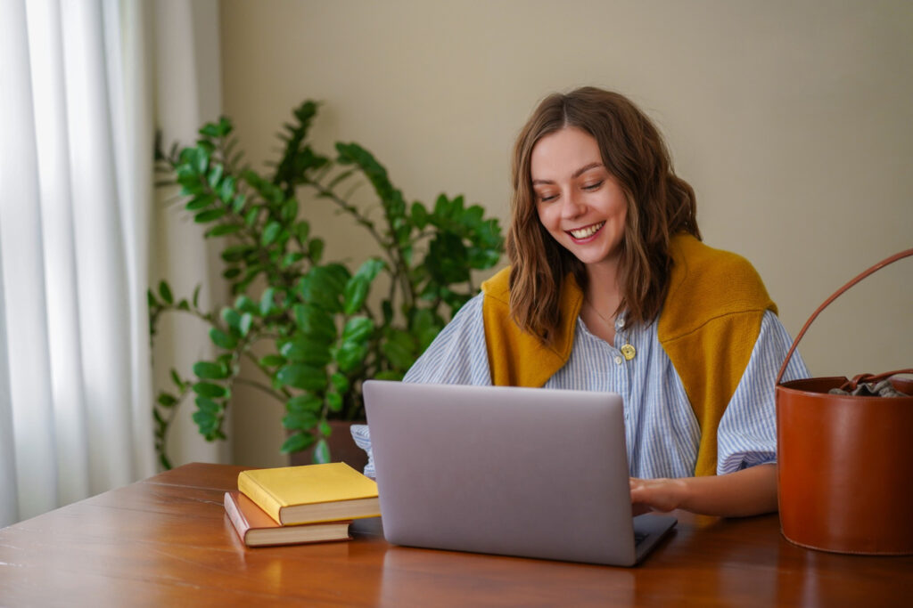 Imagem ilustrativa de mulher sorrindo diante do notebook enquanto estuda em casa, com livros sobre a mesa