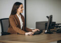 Imagem ilustrativa de recepcionista sentada à mesa de trabalho operando o computador, com impressora e telefone fixo ao fundo, em ambiente de escritório.