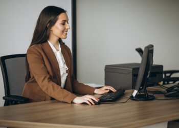 Imagem ilustrativa de recepcionista sentada à mesa de trabalho operando o computador, com impressora e telefone fixo ao fundo, em ambiente de escritório.