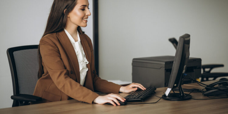 Imagem ilustrativa de recepcionista sentada à mesa de trabalho operando o computador, com impressora e telefone fixo ao fundo, em ambiente de escritório.