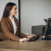 Imagem ilustrativa de recepcionista sentada à mesa de trabalho operando o computador, com impressora e telefone fixo ao fundo, em ambiente de escritório.
