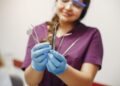 Imagem ilustrativa: profissional de jaleco roxo, óculos de proteção e luvas azuis segura instrumentais odontológicos, em referência à trilha de Auxiliar de Saúde Bucal