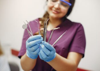 Imagem ilustrativa: profissional de jaleco roxo, óculos de proteção e luvas azuis segura instrumentais odontológicos, em referência à trilha de Auxiliar de Saúde Bucal