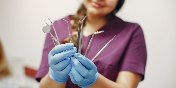 Imagem ilustrativa: profissional de jaleco roxo, óculos de proteção e luvas azuis segura instrumentais odontológicos, em referência à trilha de Auxiliar de Saúde Bucal