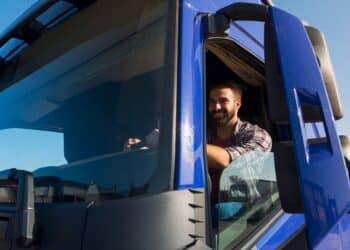 Imagem ilustrativa de motorista sorridente na cabine de um caminhão azul, com o braço apoiado na janela aberta em dia de céu limpo.