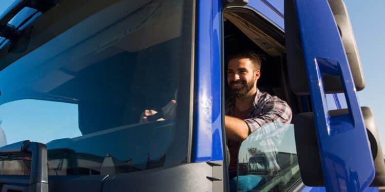 Imagem ilustrativa de motorista sorridente na cabine de um caminhão azul, com o braço apoiado na janela aberta em dia de céu limpo.