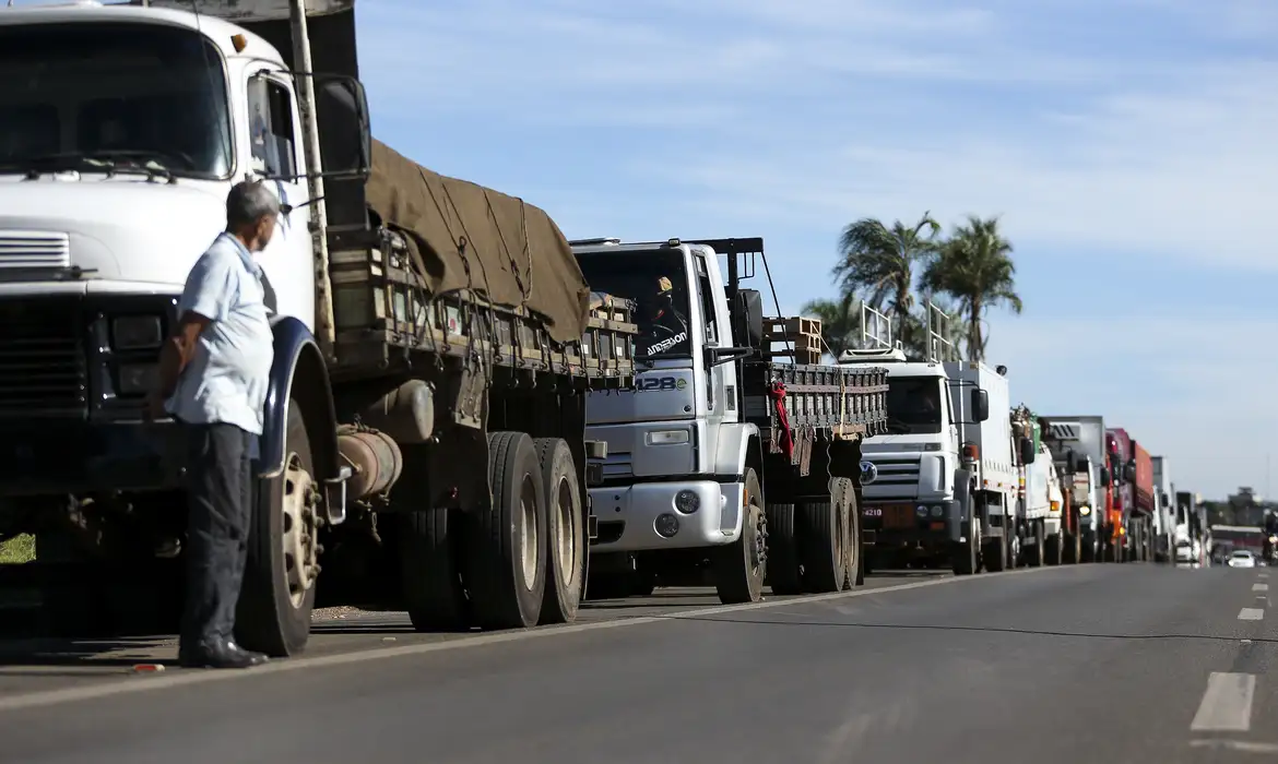 Fila de caminhões parados no acostamento de uma rodovia, com um motorista em pé ao lado da cabine observando a via.