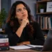 Estudante de língua portuguesa em ambiente de estudo, refletindo enquanto consulta materiais de gramática e escrita em uma mesa com livros.