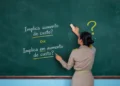 Mulher de costas escreve em quadro negro as frases "Implica aumento de custo?" e "Implica em aumento de custo?"