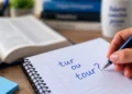 Mão escrevendo em um caderno a dúvida de português sobre o uso correto das palavras “tur” ou “tour”.