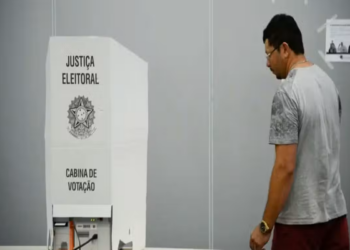 Homem adulto caminha até cabine de votação da Justiça Eleitoral do Brasil em ambiente interno, simbolizando a folga relacionada às eleições.