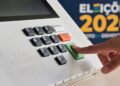 Dedo pressionando a tecla "confirma" de uma urna eletrônica, com o painel numérico em destaque e a marca Eleições 2026 ao fundo.