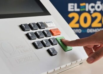 Dedo pressionando a tecla "confirma" de uma urna eletrônica, com o painel numérico em destaque e a marca Eleições 2026 ao fundo.