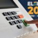 Dedo pressionando a tecla "confirma" de uma urna eletrônica, com o painel numérico em destaque e a marca Eleições 2026 ao fundo.