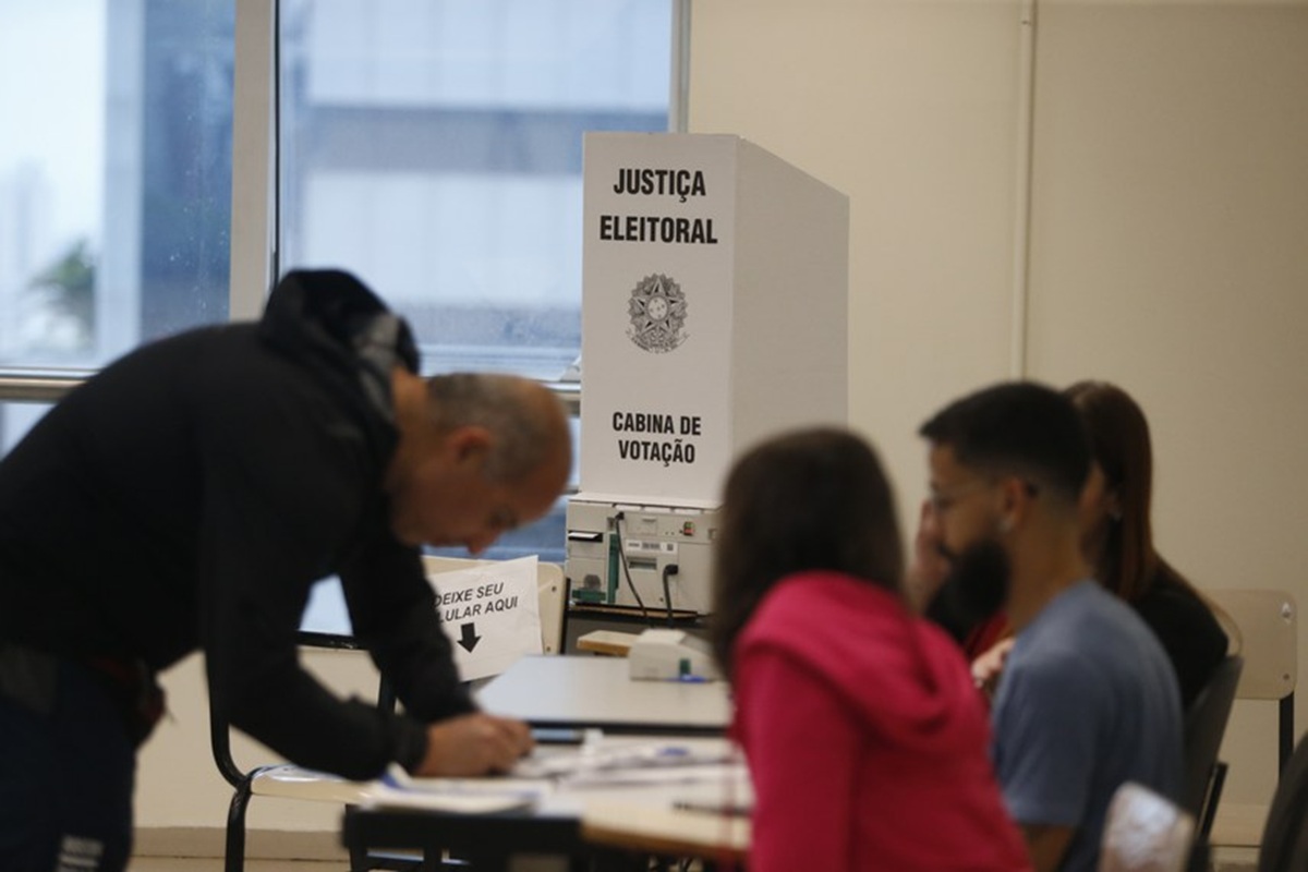 Eleitor assinando a lista de presença em uma mesa receptora de votos, ao lado da cabine de votação da Justiça Eleitoral e dos mesários, em dia de votação.