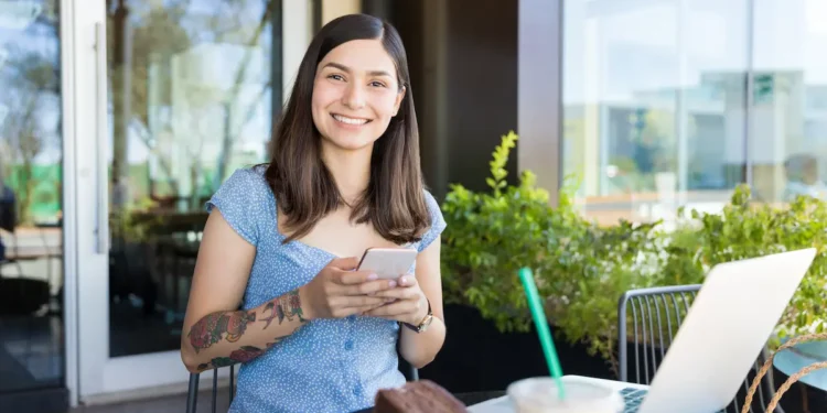 Mulher com celular ao lado de notebook representa acesso ao Fies Empreendedor para contratação de crédito.