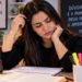 Estudante com expressão de dúvida observa o caderno, representando a incerteza sobre a grafia correta de “contraindicação”.