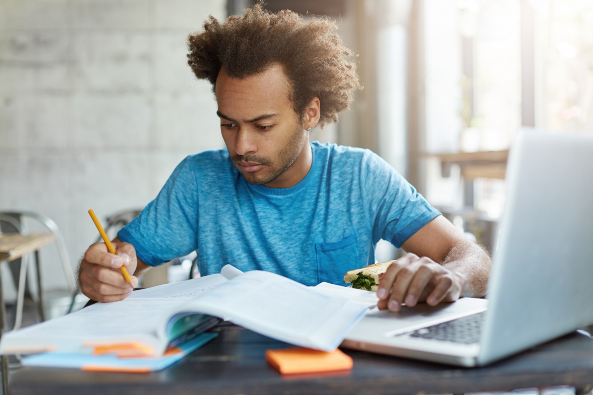Imagem ilustrativa de jovem estudando com livro aberto, lápis na mão e notebook sobre a mesa, representando quem consulta o guia do Enem