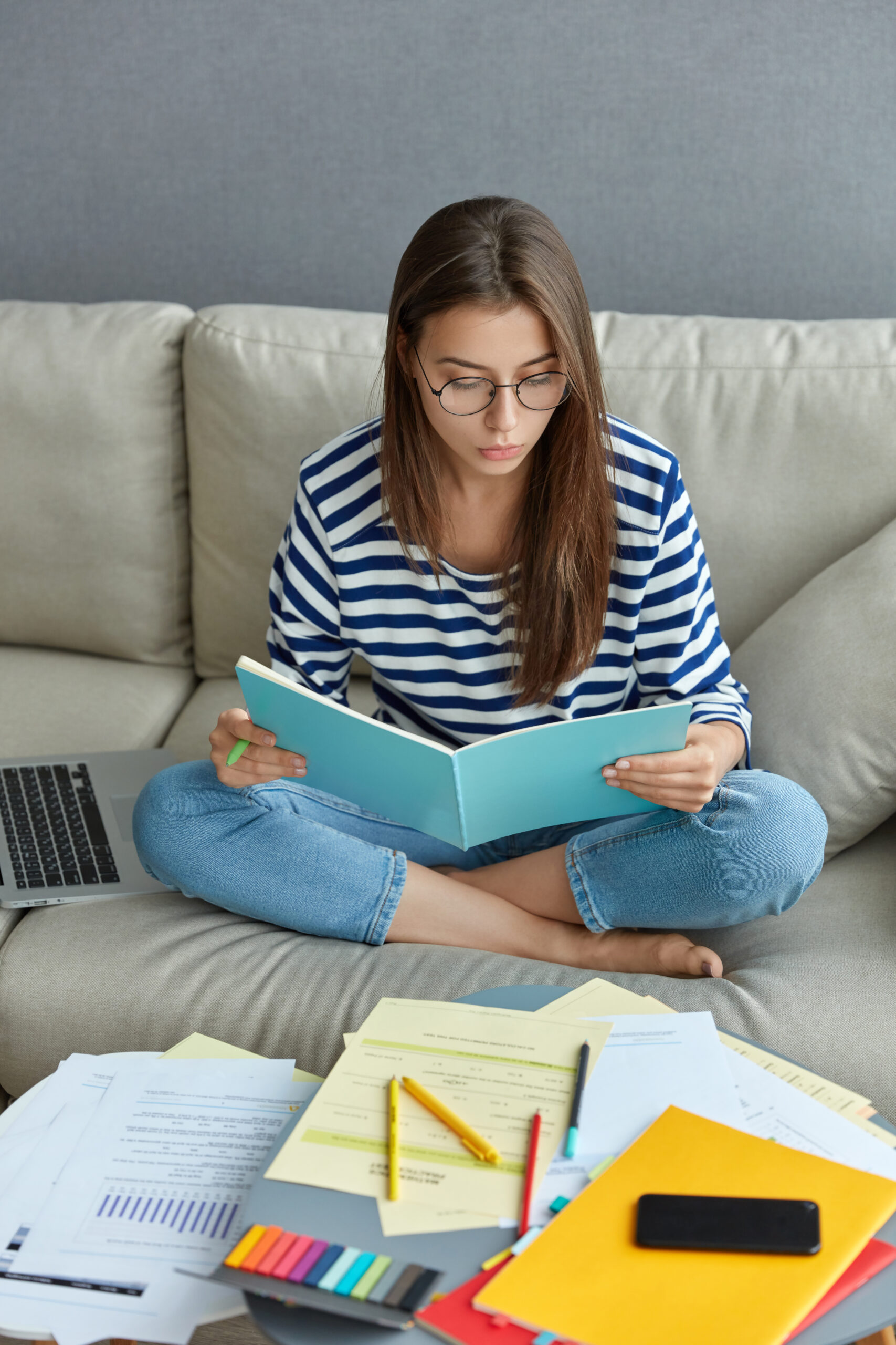 Estudante sentada no sofá lendo caderno azul rodeada de papéis, laptop e materiais coloridos, simbolizando a PND 2026.