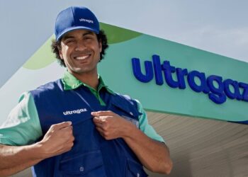 Colaborador sorridente com boné e colete da Ultragaz em frente a uma unidade da empresa, com o letreiro da marca ao fundo.