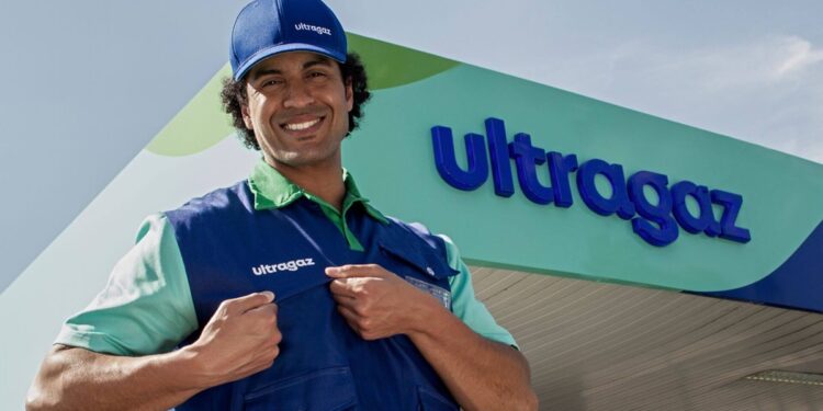 Colaborador sorridente com boné e colete da Ultragaz em frente a uma unidade da empresa, com o letreiro da marca ao fundo.
