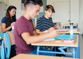 Imagem ilustrativa de estudantes em carteiras de sala de aula, com cadernos e material escolar sobre as mesas