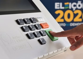 Imagem ilustrativa de dedo pressionando a tecla verde de confirmação de uma urna eletrônica, com painel das Eleições 2026 ao fundo, em referência a justificar a ausência nas Eleições 2026
