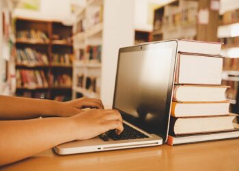 Mãos digitando em notebook com pilha de livros sobre mesa em biblioteca iluminada, simbolizando a PND 2026.