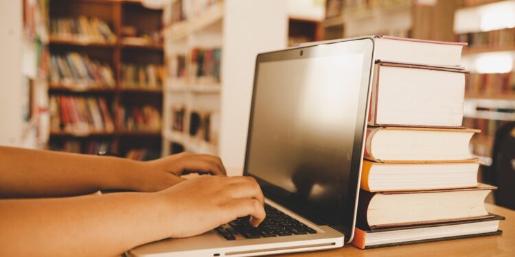 Mãos digitando em notebook com pilha de livros sobre mesa em biblioteca iluminada, simbolizando a PND 2026.