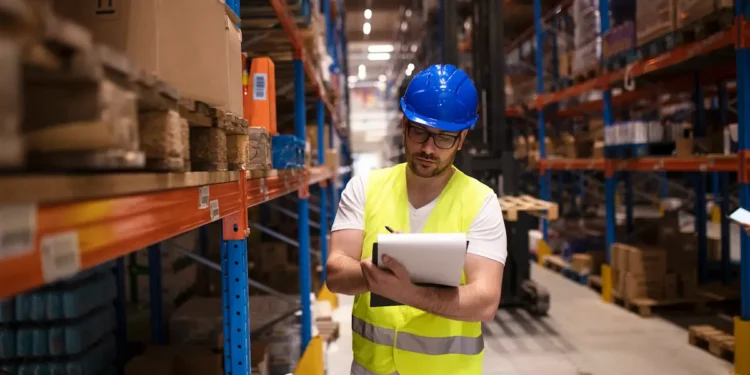 Profissional de logística em armazém realizando controle de estoque com prancheta entre corredores de produtos armazenados.