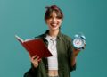 Jovem mulher sorridente segura livro vermelho aberto e relógio azul, sugerindo a escolha de horário ideal para estudar.