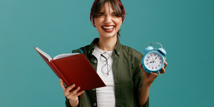 Jovem mulher sorridente segura livro vermelho aberto e relógio azul, sugerindo a escolha de horário ideal para estudar.