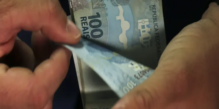 Mãos contando notas de 100 reais com destaque para cédulas azuis no centro da imagem, simbolizando o pagamento do Pe-de-Meia de setembro, que terá início no dia 21.