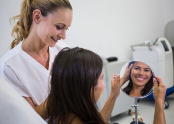 Imagem ilustrativa de esteticista de jaleco branco acompanhando o resultado do procedimento enquanto a cliente se observa em um espelho de mesa, em sala de atendimento.