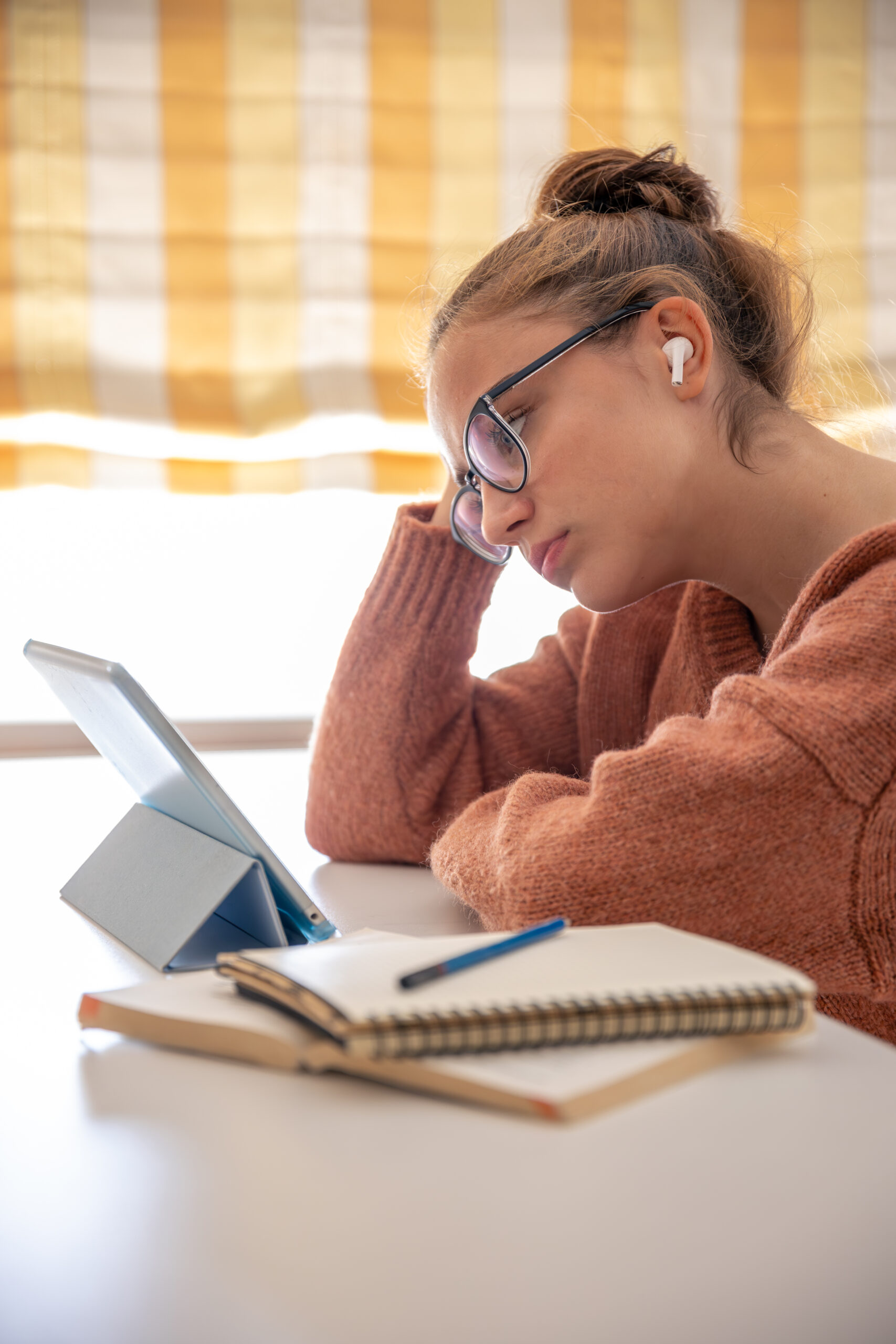 Estudante com óculos e fone sem fio no ouvido, estudando com tablet apoiado na mesa, caderno espiral aberto e caneta ao lado, apoiando o rosto na mão em postura de concentração, simbolizando quais elementos considerar antes de montar um plano de estudos para o Enem.