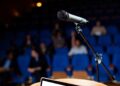 Microfone em pedestal posicionado em palco com plateia desfocada e poltronas azuis ao fundo, simbolizando o plural de porta-voz.
