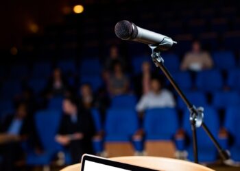 Microfone em pedestal posicionado em palco com plateia desfocada e poltronas azuis ao fundo, simbolizando o plural de porta-voz.