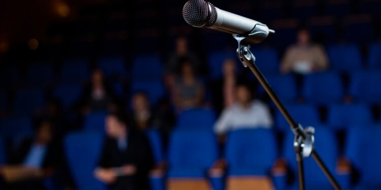 Microfone em pedestal posicionado em palco com plateia desfocada e poltronas azuis ao fundo, simbolizando o plural de porta-voz.