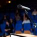Microfone em pedestal posicionado em palco com plateia desfocada e poltronas azuis ao fundo, simbolizando o plural de porta-voz.