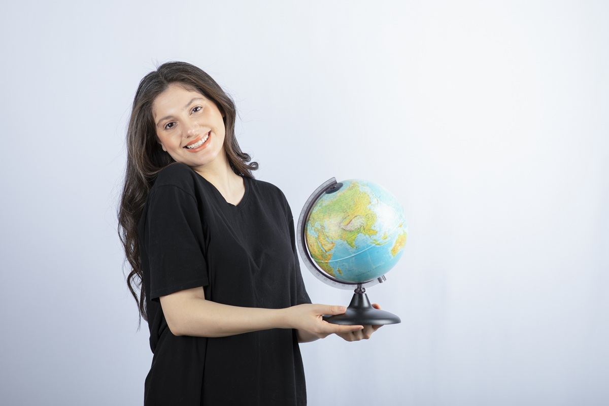 Mulher jovem sorridente veste camiseta preta e segura globo terrestre com as duas mãos, simbolizando a remuneração do professor de Geografia.